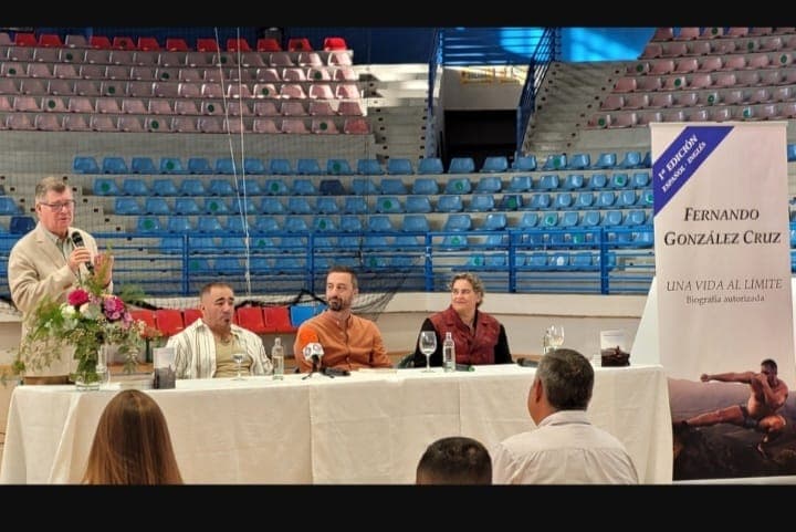 Mesa de la presentación de «Una vida al límite» en un pabellón deportivo: cuatro personas sentadas, una de ellas hablando por el micrófono, y a la derecha el cartel vertical del libro